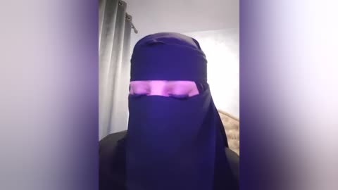 Media: A video of a person wearing a black niqab with a glowing pink visor, creating a surreal, otherworldly effect. The background features a neutral-colored room with gray curtains.