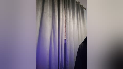 Media: Video of a room with a large, light blue and gray curtain drawn partially to the side, revealing a white wall with a small window. A black leather jacket hangs on a coat rack in the corner.