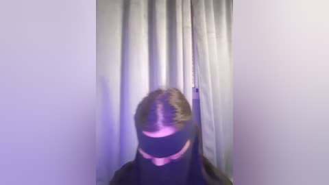 Media: Video of a person wearing a black hooded mask with a large white triangle, standing in front of a white curtain. The image is blurry and low quality, making it difficult to discern details about the person's gender or age.