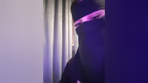 Media: Video of a person partially obscured by a black mask, with purple and pink lighting highlighting their face, in a dimly lit room with grey curtains.