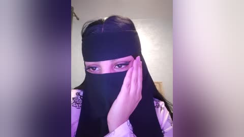 Media: Video of a woman with long black hair, wearing a black hijab, and a white shirt with floral embroidery, covering her mouth with her hand, standing in a dimly lit hallway.