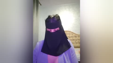 Media: A video of a person wearing a black burqa with a pink strip over the eyes, standing in a beige-tiled room with a wooden headboard and a partially visible bed.