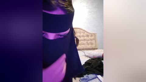 Media: A video of a woman in a black dress standing beside a bed with scattered clothes, featuring a headboard with a beige pattern and a purple light illuminating the scene.