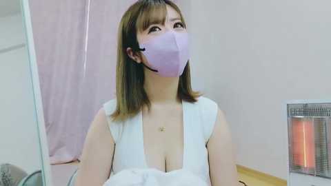 Media: Video of an East Asian woman with shoulder-length brown hair, wearing a lavender face mask and white sleeveless top, sitting indoors.