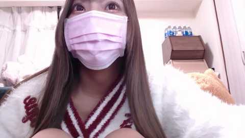 Media: Video of an Asian woman with long brown hair, wearing a white and red striped kimono, pink surgical mask, and a white fur coat, in a cluttered bedroom with a bed, stuffed animal, and water bottles on a table.