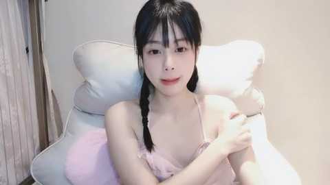 Media: A video of an East Asian woman with fair skin and black hair in pigtails, wearing a pink spaghetti-strap camisole, lying on a white pillow, against a plain beige wall, smiling softly.