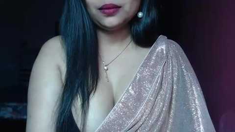 Media: A video of a woman with long, straight black hair, fair skin, and wearing a glittering silver saree with a deep neckline, accentuated by a pearl necklace.