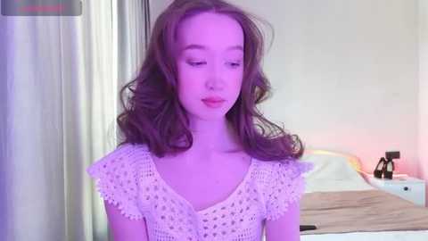 Media: A video of a young woman with fair skin and wavy, shoulder-length brown hair, wearing a light pink crochet top. She stands in a softly lit bedroom with a bed and white curtains in the background.