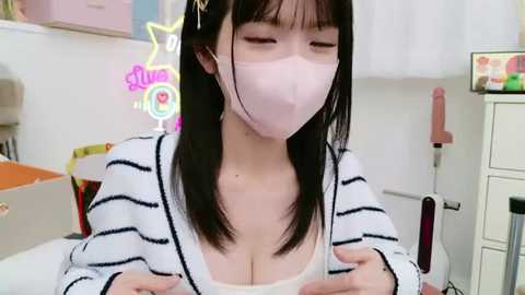 Media: Video of an East Asian woman with straight black hair and a pink mask, wearing a white and black striped sweater, seated in a bright, modern room with neon decor.