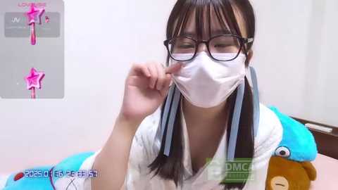 Media: Video of an Asian woman in a white lab coat with a blue collar, wearing a white face mask and black-framed glasses, examining a small creature in a laboratory setting.