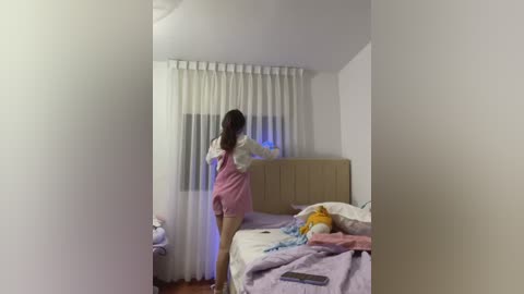 Video of a young woman in a pink dress standing in a dimly lit bedroom with a messy bed and white curtains.