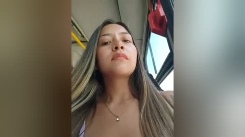 Media: A video of a young woman with long, straight, dark brown hair, wearing a white top, sitting inside a car. She has a neutral expression and a small gold necklace. The background shows a blurred interior with red clothing hanging.