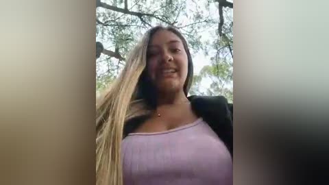 Video of a smiling, light-skinned woman with long, straight blonde hair, wearing a purple tank top and black jacket, standing outdoors against a backdrop of green trees.