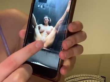 Media: A video shows a woman with fair skin and medium breasts, nude, with legs spread, on a smartphone screen. Her hand holds the phone, and the background features a bathroom with a tiled wall.