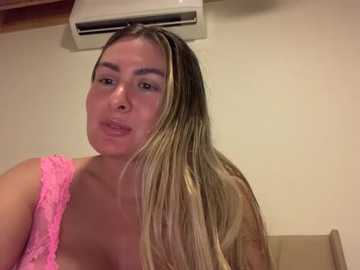 Media: Video of a light-skinned woman with long, straight, blonde hair, wearing a pink lace top. She is indoors with beige walls, an air conditioner mounted above her, and a beige chair visible in the background.