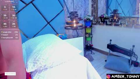 Media: Video of a modern bedroom with a white bed, turquoise headboard, and blue geometric-patterned walls. Two pendant lights hang above, a massage chair rests in the corner, and a potted plant decorates the space.