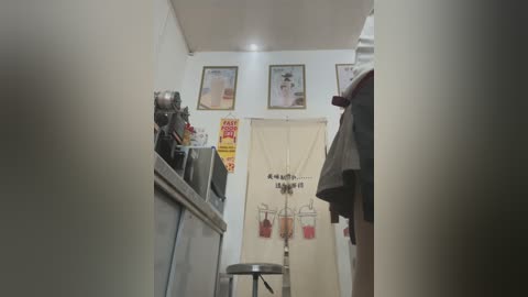 A video of a narrow, cluttered room with a white ceiling, beige walls, and a small table. A metal stool is in the foreground. Vintage posters and a coffee machine are on the left, while a coat hangs on the right.