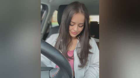 Media: Video of a young woman with long brown hair, wearing a pink shirt and light jacket, sitting in a car, focused on the steering wheel.