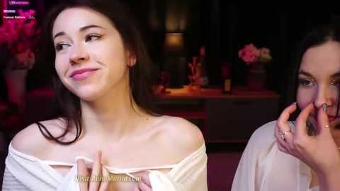 Media: Video of two women in a dimly lit room: one with light skin, dark hair, and a white top, smiling, and the other with dark hair, holding a phone, in a white top.