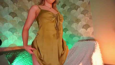 Media: Video of a fair-skinned woman with long blonde hair wearing a mustard-yellow satin slip dress, posing indoors with a textured, patterned wallpaper and a green-lit room.