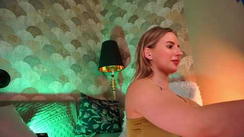 Media: Video of a fair-skinned, blonde woman with medium-sized breasts in a mustard-yellow top, sitting on a bed with green and gold patterned pillows, under a green and beige wavy wallpaper, with a black lampshade and a green blanket in the background.