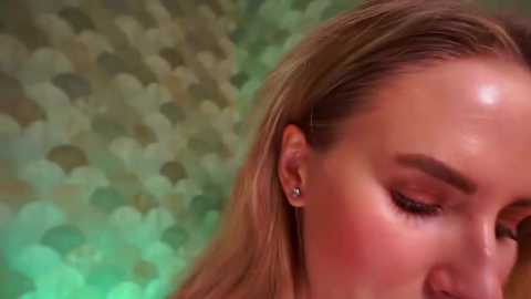 Video of a close-up of a woman with fair skin, long blonde hair, and closed eyes. She has subtle makeup, wearing small stud earrings. The background is a blurred, green and beige abstract pattern.