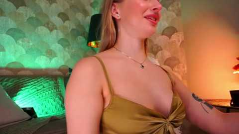 Media: Video of a smiling woman with blonde hair, wearing a low-cut olive green dress, standing in a dimly lit bedroom with floral wallpaper and a green-lit lamp.