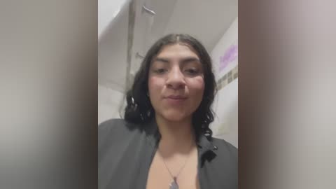 Media: Video of a young woman with medium brown skin, shoulder-length black hair, wearing a black shirt, standing in a bathroom with white tiles and a shower curtain.