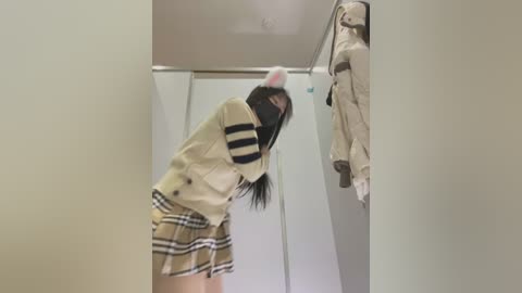 Media: Video of a young woman in a beige jacket, black mask, and plaid skirt, bending over in a changing room, with a beige coat hanging on a hook.