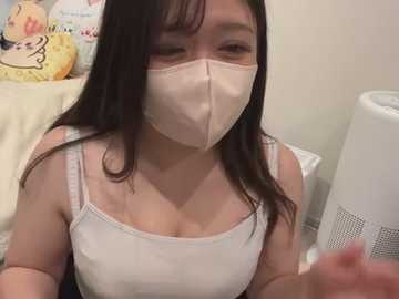 Media: Video of an East Asian woman with long black hair, wearing a white mask, white tank top, and sitting on a bed with plush toys and a white air purifier in the background.