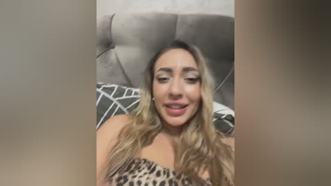 Media: A video of a light-skinned woman with long, wavy blonde hair, wearing a leopard-print top, lying on a gray couch with a geometric-patterned pillow, smiling.