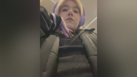 Media: Video of a young woman with blonde hair, wearing purple headphones and a striped sweater, sitting in a dimly lit room, leaning forward, with a slightly blurred background.