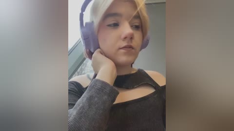 Video of a young woman with light skin, short blonde hair, wearing purple over-ear headphones and a black off-shoulder top, leaning against a window with blurred background.