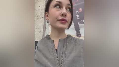 Media: A video of a young woman with fair skin and short dark hair, wearing a grey zip-up jacket, looking contemplative against a white brick wall with a poster in the background.