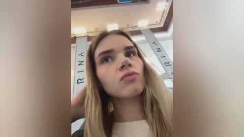 A video of a young woman with long, straight blonde hair, wearing a white top, standing in a modern, brightly lit room with a ceiling featuring white panels and a blue light fixture.