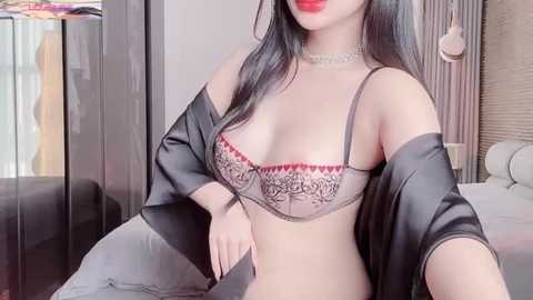 Media: Video of an Asian woman with long black hair, wearing a sheer lace bra with red accents, black robe, and choker, sitting on a bed in a softly lit room.