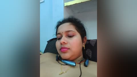 Media: Video of a woman with medium brown skin and dark hair resting her head on a cushion, wearing a beige top, gold necklace, and blue headphones.