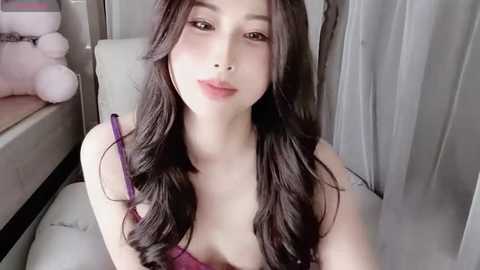 Media: A video of a young Asian woman with long, dark hair, fair skin, and a slender physique, wearing a purple top, sitting on a white couch with a teddy bear nearby.