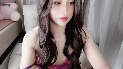 Media: Video of a young Asian woman with long, wavy black hair, wearing a pink lace bra, sitting on a beige leather couch in a softly lit room with light curtains.