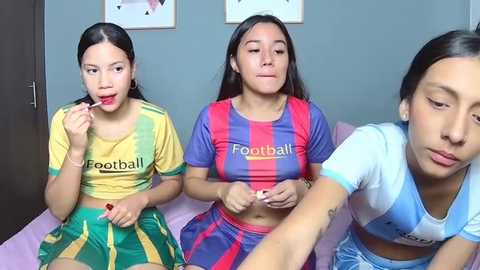 Video of three young Latina women with long dark hair, wearing soccer jerseys and skirts, sitting on a bed, eating candy, in a brightly lit room with blue walls and framed art.