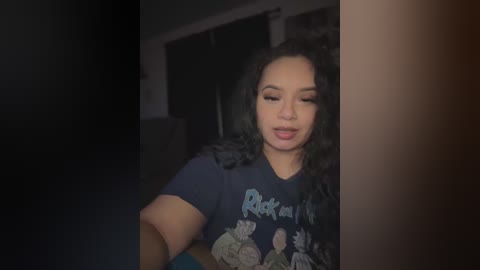 Video of a smiling, light-skinned Black woman with curly hair, wearing a black graphic tee, in a dimly-lit room with dark curtains.