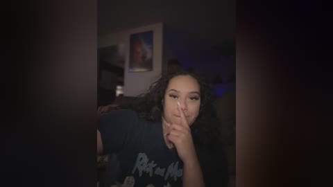 Media: Video of a young woman with curly hair, dark skin, and a large nose, wearing a dark T-shirt, holding her finger to her lips, in a dimly lit room with a framed poster on the wall.