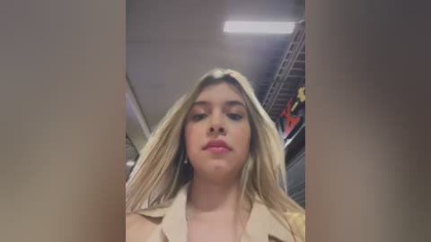 Media: Video of a blonde woman with fair skin, wearing a beige trench coat, looking downwards with a neutral expression. Background features industrial elements and a ceiling light.