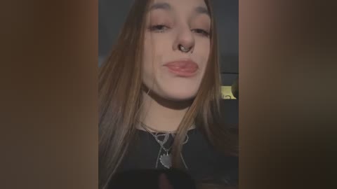 Media: Video of a young woman with long brown hair, fair skin, and a nose ring, puckering her lips in a pout. She wears a black top and layered necklaces.