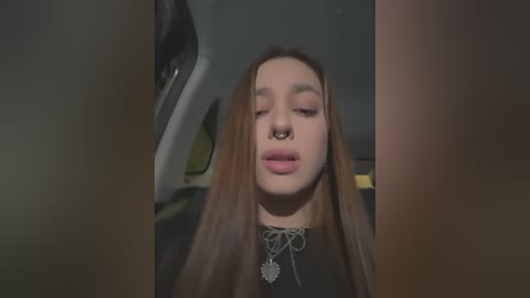 Media: Video of a young woman with long brown hair, pale skin, and full lips, wearing a black top and multiple necklaces, inside a car at night.