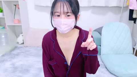 Media: Video of a young Asian woman with light skin, wearing a pink face mask and maroon pajamas, with a bed and white curtains in the background.