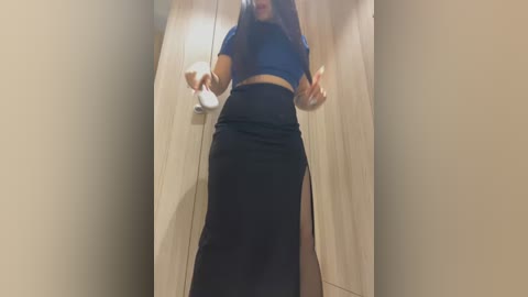 Media: Video of a young woman with long, dark hair, wearing a navy blue crop top and high-waisted black skirt with a slit, standing in a beige hallway, making a peace sign.