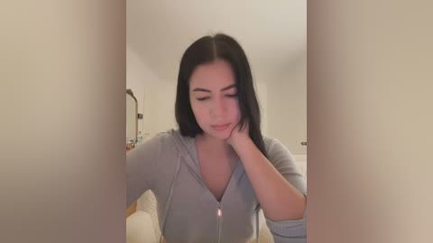 Media: Video of a young woman with long black hair, light skin, and a slim build, wearing a gray hoodie, resting her head on her hand in a soft, intimate indoor setting.