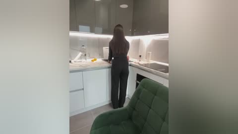 Media: Video of a modern kitchen with a woman in a black dress facing the counter. The kitchen has sleek white cabinets, a green chair, and a minimalist design.