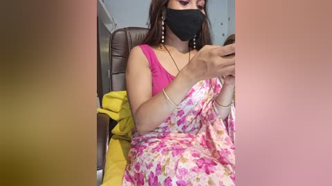 Media: A video shows a woman with medium skin tone, wearing a pink floral sari, black face mask, and gold earrings, seated on a yellow cushion in a bus.